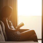 Frau liest Buch auf Couch vor geöffneter Balkontür, die Sonne scheint herein